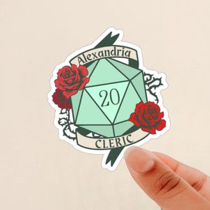 May include: A green twenty-sided die with the number 20 on it, surrounded by red roses and thorns. A banner above the die reads "Alexandria Cleric".