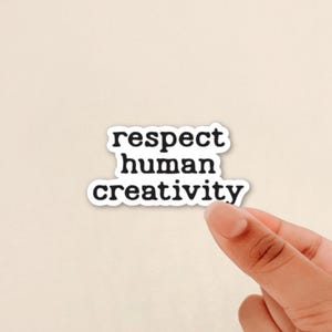 May include: A white sticker with the words "respect human creativity" in a black, bold font. The sticker is held by a person's hand against a light beige background.
