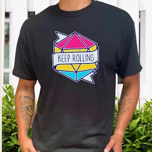May include: Black t-shirt featuring a geometric design in pink, yellow, and blue, with the words "KEEP ROLLING" on a banner. The design is outlined in blue. The shirt is a casual wear item.