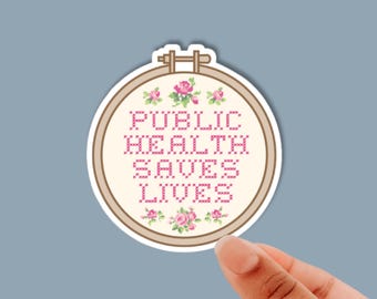 May include: A circular sticker featuring a cross-stitch design inside a wooden embroidery hoop. The cream-colored fabric displays the pink text PUBLIC HEALTH SAVES LIVES, surrounded by small pink and green floral motifs at the top and bottom.