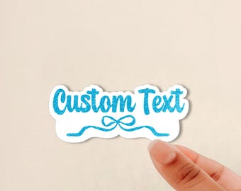 May include: A die-cut sticker featuring the words Custom Text in a bright blue glitter finish. Below the text is a stylized blue glitter ribbon bow graphic with two flowing tails extending outward to the left and right sides of the sticker.