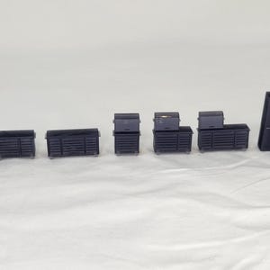 Puede incluir: Una colección de miniaturas de cofres y armarios de herramientas negros de varios tamaños, dispuestos en fila. Los cofres tienen un diseño acanalado y algunos tienen cajones. La pieza más grande se asemeja a una estación de trabajo con una ventana.