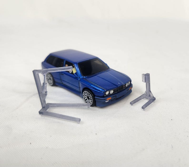May include: A blue toy car with silver rims and a clear plastic car lift. The car lift is in two pieces. The car is a station wagon style. The background is white.