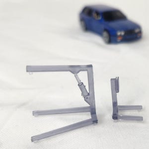 May include: Two transparent plastic model car lift components are displayed, with a blue toy car in the background. The lift parts are gray and have a detailed design, suggesting a miniature automotive theme.