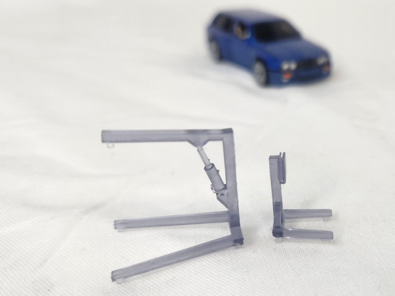 May include: Two transparent plastic model car lift components are displayed, with a blue toy car in the background. The lift parts are gray and have a detailed design, suggesting a miniature automotive theme.