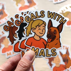 May include: A collection of white stickers featuring the text "Gals With Animals". Each sticker shows a cartoon illustration of a woman with a blonde ponytail and a German Shepherd dog. The woman is wearing an orange top.