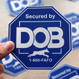 May include: Blue octagonal sticker with white text reading "Secured by DOB" and a running dog silhouette. The sticker also includes the phone number 1-800-FAFO. Several similar stickers are visible in the background.