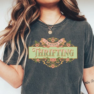 May include: A gray t-shirt with the text "I'd rather be thrifting" in a vintage-inspired design with floral accents. The text is in a light green banner with gold trim.