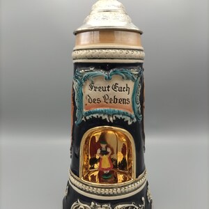 May include: A ceramic beer stein with a silver lid and a musical mechanism. The stein features a blue and white design with a scene of a woman in traditional German dress. The text "Freut Euch des Lebens" is written on the stein.