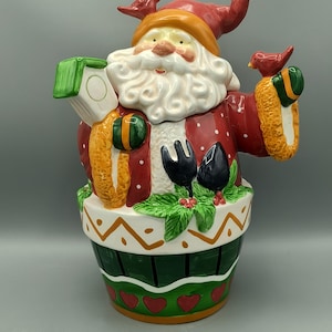 May include: A ceramic Santa Claus cookie jar. The jar is red with white polka dots and a white beard. Santa is wearing a red hat with a gold star and holding a birdhouse and a bird. The base is green with a white and orange design.