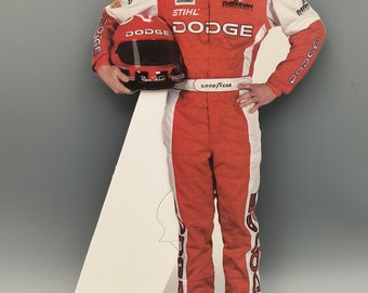May include: A cardboard cutout of a racing driver wearing a red racing suit with white accents. The driver is holding a helmet and has a serious expression on his face. The suit has the words "Dodge" and "Good Year" printed on it. The driver is standing on a white cardboard base.