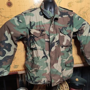 May include: A green, brown, and black camouflage jacket with a button-up front and two large pockets on the front.