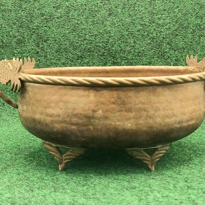 May include: A bronze-colored decorative bowl with three ornate feet and two handles. The bowl has a rope-like trim around the rim and decorative elements on the handles. The bowl is set against a green background.