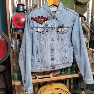 May include: A light blue denim jacket with a red heart-shaped patch and other small patches. The jacket has a button closure and two front pockets.