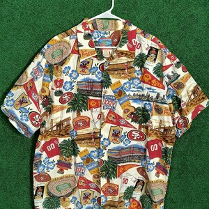 May include: Short-sleeved button-down shirt with a colorful print featuring the San Francisco 49ers football team. The design includes team logos, stadium illustrations, and the words "Super Bowl" and "San Francisco". The shirt is on a white hanger.