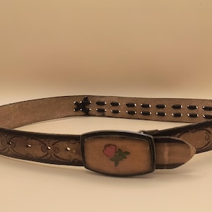 May include: A brown leather belt with a metal buckle featuring a pink rose design. The belt has a decorative stitched pattern and a row of holes for adjusting the size.