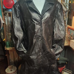 May include: Black leather jacket with a patchwork design. The jacket has a collared neckline, three buttons, and two front pockets. The jacket is displayed on a mannequin.