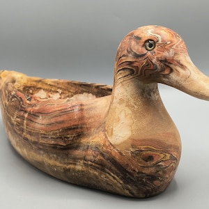 Puede incluir: Una maceta decorativa con forma de pato y aspecto marmolado natural. La maceta de cerámica presenta tonos marrones, beiges y rojizos, con un cuerpo ahuecado para plantas u otros objetos. El pato tiene una cabeza y un pico detallados.