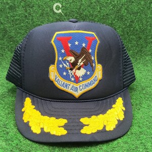 May include: Navy blue trucker hat with a mesh back and a curved brim. The front features an embroidered patch with an eagle, lightning bolt, and the text "Valiant Air Command". Yellow leaf designs adorn the brim.