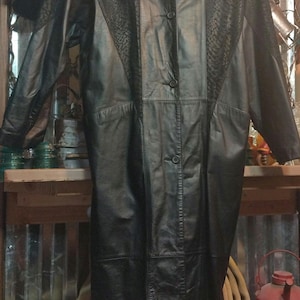 May include: A long black leather coat with a button-up front and a textured pattern. The coat has a collar and long sleeves.