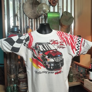 May include: White t-shirt featuring a race car design with the text "Team Auto Tire Racing" and the number 26. The shirt has a checkered flag design on the sleeve and red accents.