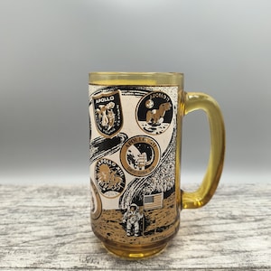 May include: A yellow glass mug with a black and white design featuring the Apollo missions. The mug has a handle and a round base. The design includes the Apollo 11, 12, and 13 mission patches, as well as images of astronauts on the moon.
