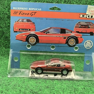 May include: A die-cast metal replica of an '88 Fiero GT car. The red model is displayed in a clear plastic case on a blue cardboard backing with illustrations of the car. The packaging reads "Blueprint Replicas" and "ERTL".