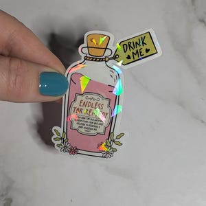 May include: A holographic sticker of a pink potion bottle with a label that reads "Endless TBR Remedy". The sticker also includes a tag that says "Drink Me" with a heart.
