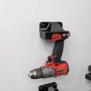 May include: A red and black cordless drill mounted on a black wall-mounted holder. The drill is positioned against a light gray wall. The holder is designed to securely store the drill.