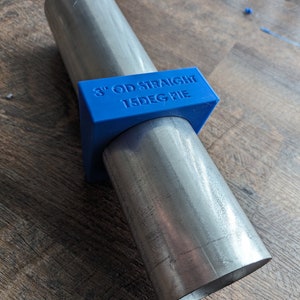 May include: A metal pipe assembly with a blue bracket. The bracket is labeled "3" OD STRAIGHT 15DEG PIE". The pipes are silver and angled. The assembly is on a wooden surface.