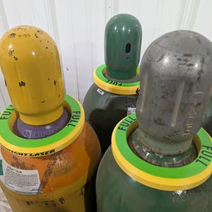 May include: A cluster of industrial gas cylinders, each with a rounded top and a green and yellow collar indicating they are full. The cylinders are various colors, including yellow, green, and silver. Some cylinders have labels with text.