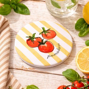 May include: Round white and yellow striped coaster with three red tomato illustrations and the word "pomodori" in cursive. The coaster is surrounded by fresh basil, lemons, and a glass of water.