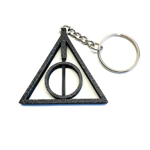 Puede incluir: Llavero de metal negro con el símbolo de las Reliquias de la Muerte de la serie Harry Potter. El símbolo es un triángulo con un círculo en el interior y una línea vertical que atraviesa el círculo.