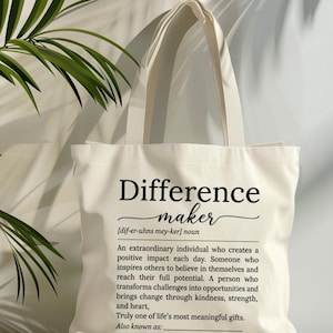 May include: A cream-coloured canvas tote bag featuring black printed text that defines the term Difference maker. The text includes a phonetic pronunciation, a noun classification, and a descriptive paragraph about creating positive impact.