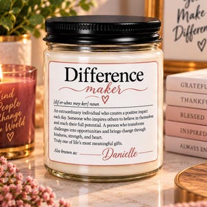 Può includere: Un barattolo di candela in vetro trasparente con coperchio nero e un'etichetta bianca che riporta il testo Difference maker, una definizione in stile dizionario e il nome Danielle. La candela contiene cera color bianco sporco.