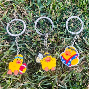 May include: Three metal keychains featuring yellow rubber ducks. The first duck wears a blue headband and holds a red and white football. The second duck wears black sunglasses and holds a beer mug. The third duck wears sunglasses and holds a surfboard.