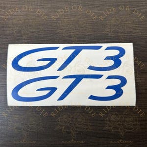 May include: A rectangular white decal sheet featuring two identical blue GT3 logos printed in a bold, stylized font. The text GT3 is displayed twice, one above the other, on a smooth white adhesive backing designed for application to surfaces.