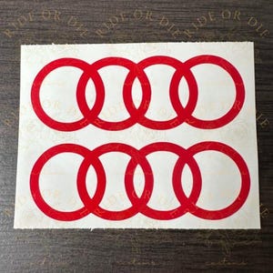 May include: Two red Audi logo decals. Each decal features two rows of four interlocking rings. The decals are on a white background with a floral pattern and the words "Ride or Die" in a circular pattern.