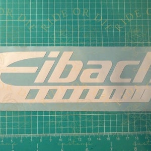 May include: White vinyl sticker with the word "Eibach" in a bold, white font. The sticker has a curved design with a series of white lines underneath the word.