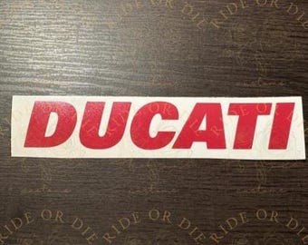 May include: A rectangular white adhesive decal featuring the word DUCATI printed in bold, red, sans-serif capital letters. The sticker has a smooth, glossy finish and is presented as a single, flat graphic element for application on surfaces.