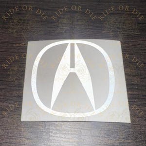 May include: A white vinyl decal featuring the Acura logo, which consists of a stylized letter A inside an oval frame. The decal has a subtle floral rose pattern integrated into the white design and is presented on a square backing sheet.