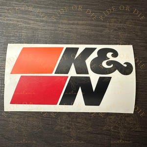 May include: A rectangular adhesive decal featuring the black text K&N positioned to the right of two stacked parallelograms. The top parallelogram is orange, and the bottom parallelogram is red. The entire design is on a white background.