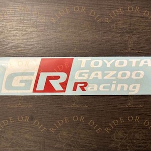 May include: A rectangular vinyl decal featuring the Toyota Gazoo Racing logo. The design includes a large white and red GR emblem on the left, followed by the white text TOYOTA GAZOO Racing on the right, all set against a clear backing.