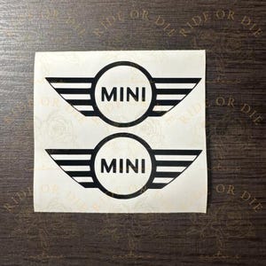 May include: Two black vinyl decals featuring the MINI logo, each consisting of a central circle with the word MINI in bold capital letters and stylized horizontal wings on either side, presented on a square white backing sheet.