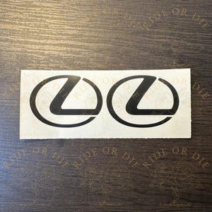 May include: Two black Lexus logo decals on a white background. The decals are in the shape of a stylized "L" inside a circle. The background has the words "RIDE OR DIE" and "customs" printed on it.