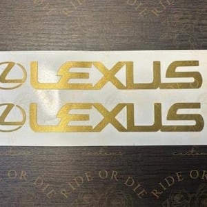 May include: Two gold-colored Lexus logo decals on a white background. The decals feature the Lexus emblem and the word "LEXUS" in a bold, sans-serif font. The decals are likely for automotive customization.