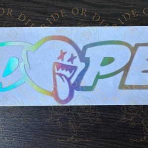 May include: A holographic vinyl sticker with the word "DOPE" in a rainbow color scheme. The "O" is a cartoon ghost with a tongue sticking out and two stars above its eyes.