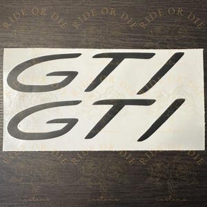 May include: A rectangular white decal sheet featuring two black GTI logos in a bold, slanted sans-serif font. The text RIDE OR DIE is repeated in a faint, watermark-style pattern across the background of the sticker sheet.