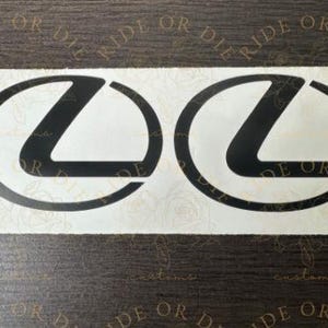 May include: Two black Lexus logo decals on a white background. The decals are oval-shaped with a stylized "L" inside. The background has a floral pattern and the words "RIDE OR DIE" and "customs" are printed on the background.