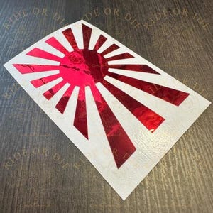 May include: A rectangular decal featuring a metallic red rising sun design with radiating beams against a white background. The design includes a central circular element with rays extending outward, creating a bold graphic pattern on a flat surface.